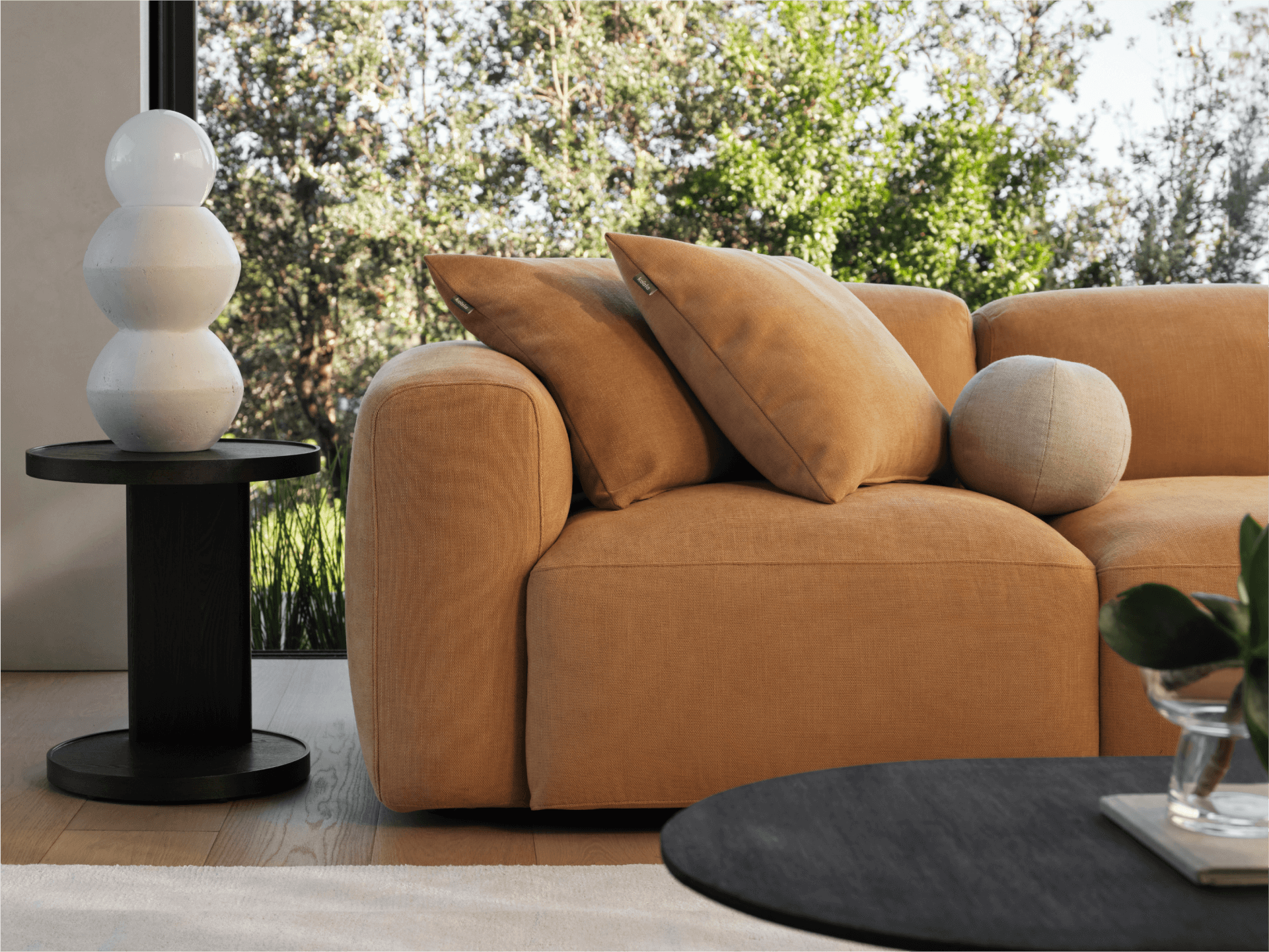 Luxurious caramel-coloured, fabric sofa with plush cushions, set in a light-filled modern living room, paired with a black round side table and ceramic lamp.