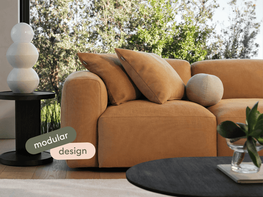 Luxurious modular sofa in tan suede with plush cushions, set in a chic living space. Black round side table with sculptural lamp complements the style.