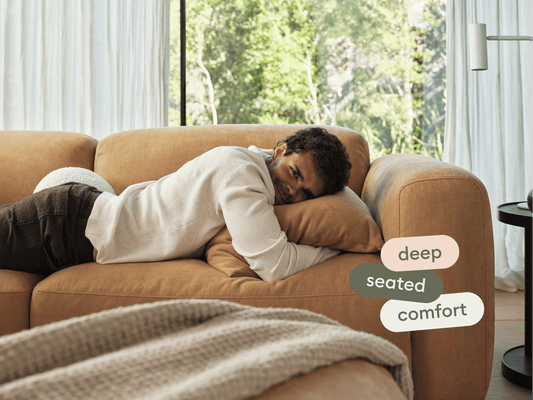 Luxury caramel leather sofa with deep seating, plush cushions, and sleek curved arms. Set in a spacious, light-filled living room with floor-to-ceiling windows.