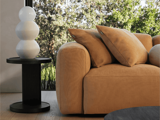 Modern side table with sleek black finish, cylindrical design, and minimalist aesthetic; set beside a plush caramel sofa in a sunlit living room.