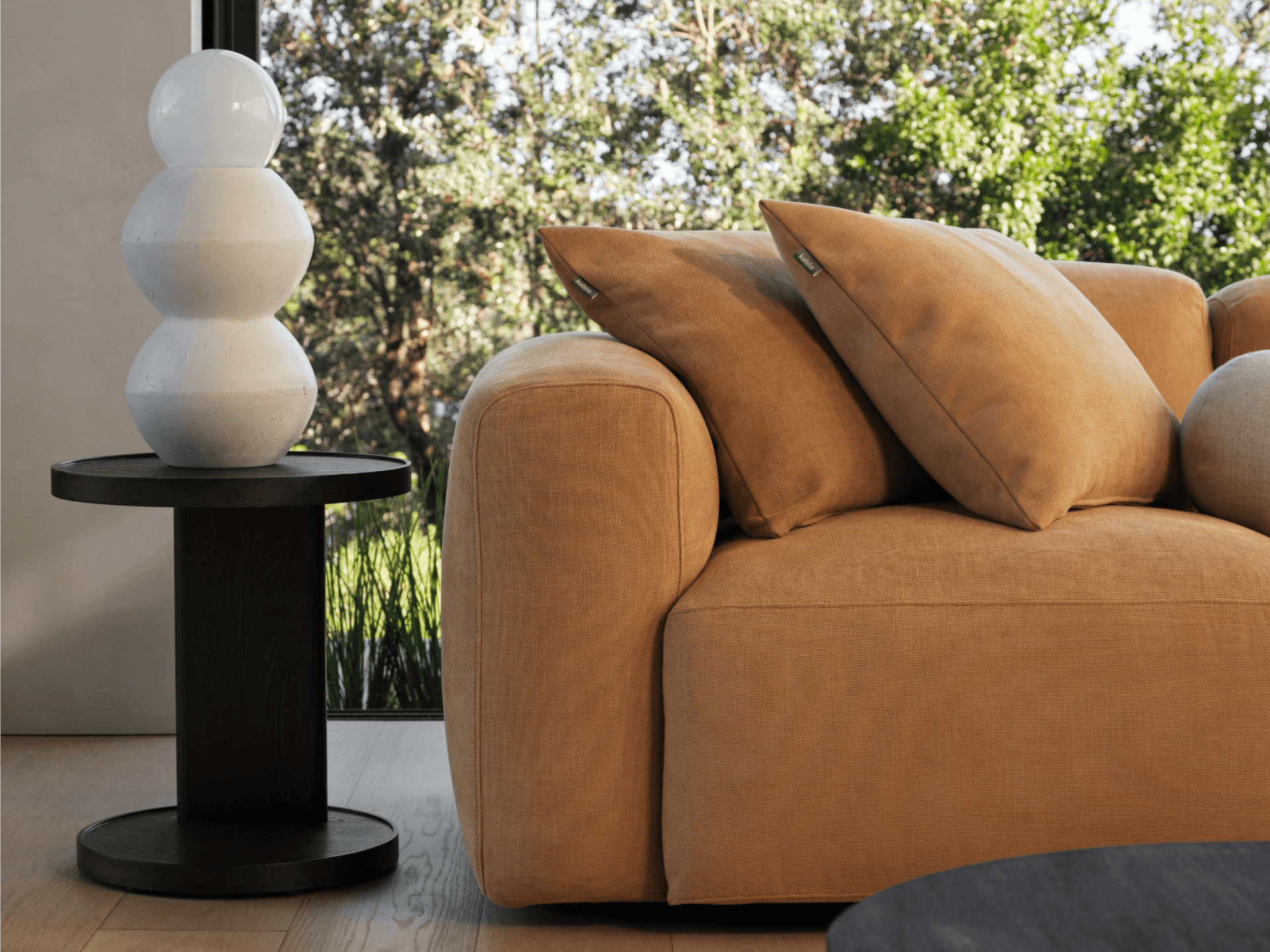 Luxurious caramel fabric sofa with plush cushions alongside a sleek black side table. Matte white ceramic lamp, natural light, and serene outdoor view.