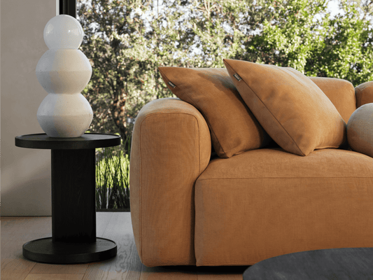 Luxurious caramel fabric sofa with plush cushions alongside a sleek black side table. Matte white ceramic lamp, natural light, and serene outdoor view.