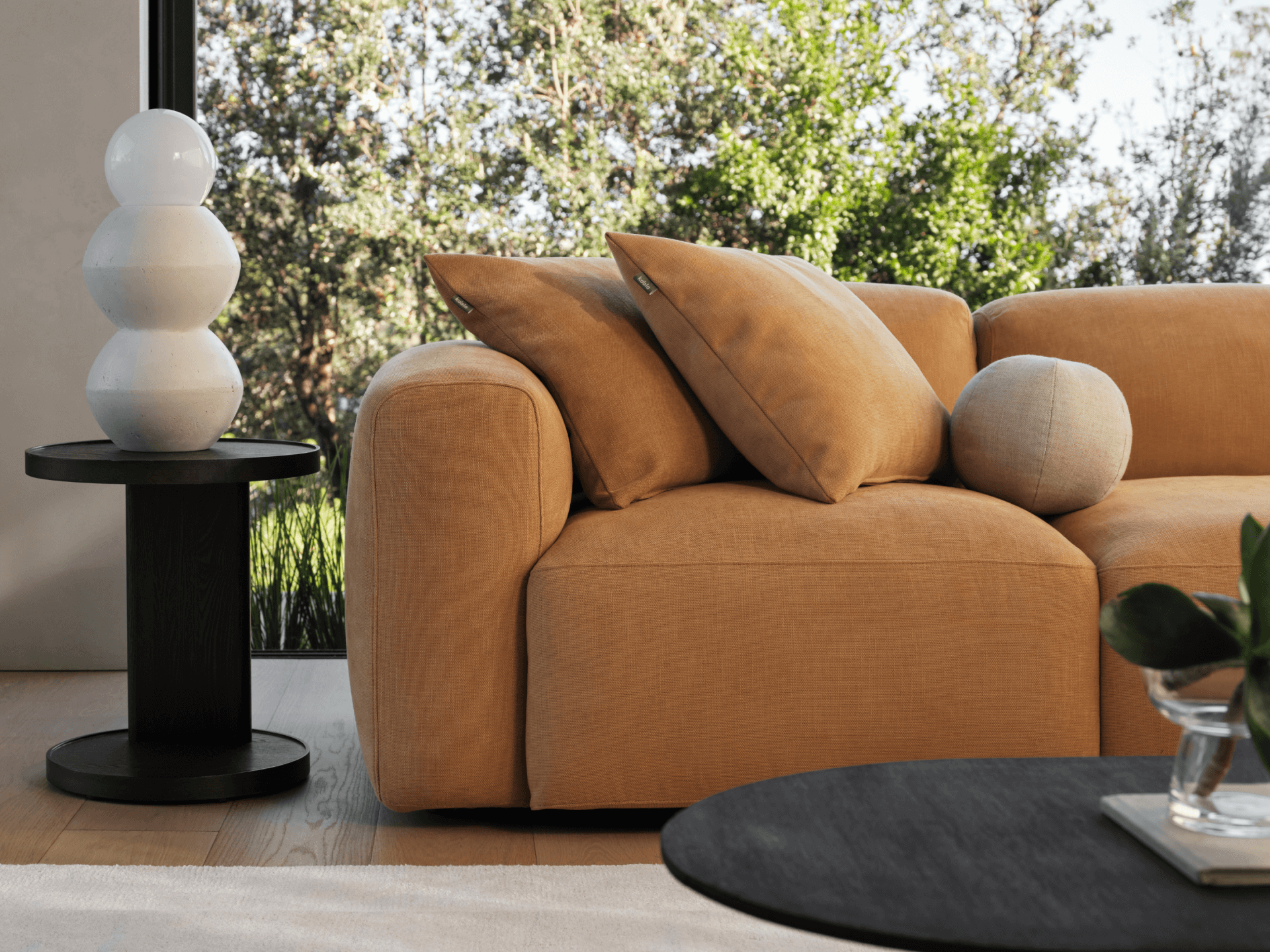 Luxury caramel velvet sofa with plush cushions, featuring rounded armrests. Set on light wooden flooring, accompanied by a round black side table.
