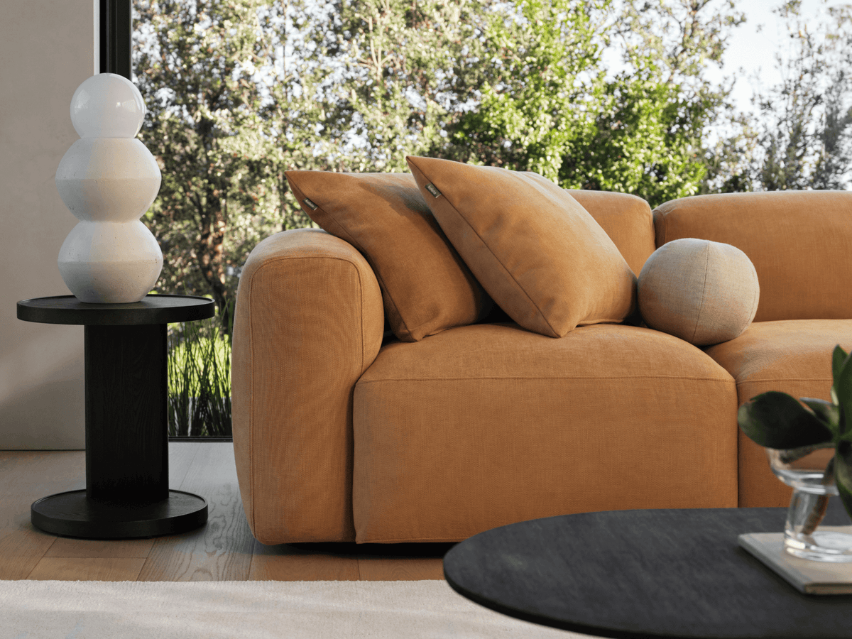 Luxurious mustard sofa with plush fabric, squared design, and deep cushions in a light-filled modern living room. Features black side table and greenery backdrop.
