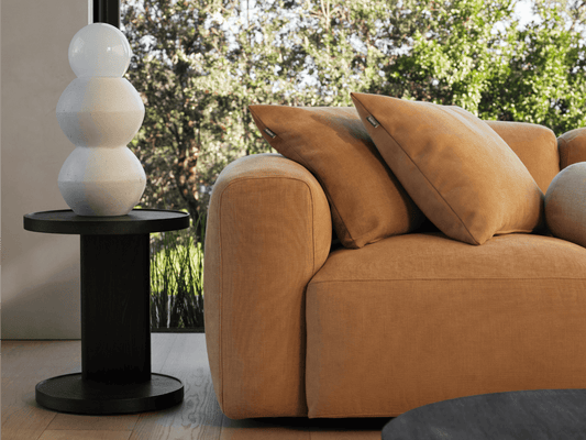 Luxurious tan fabric sofa with plush cushions, paired with a modern black wood side table. Set in a sunlit room overlooking lush greenery.