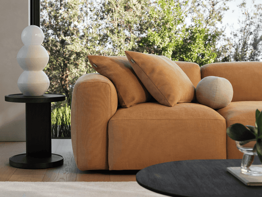 Luxurious beige velvet sofa with plush cushions, set in a sunlit room. Accompanied by a sleek black side table with a modern ceramic lamp.