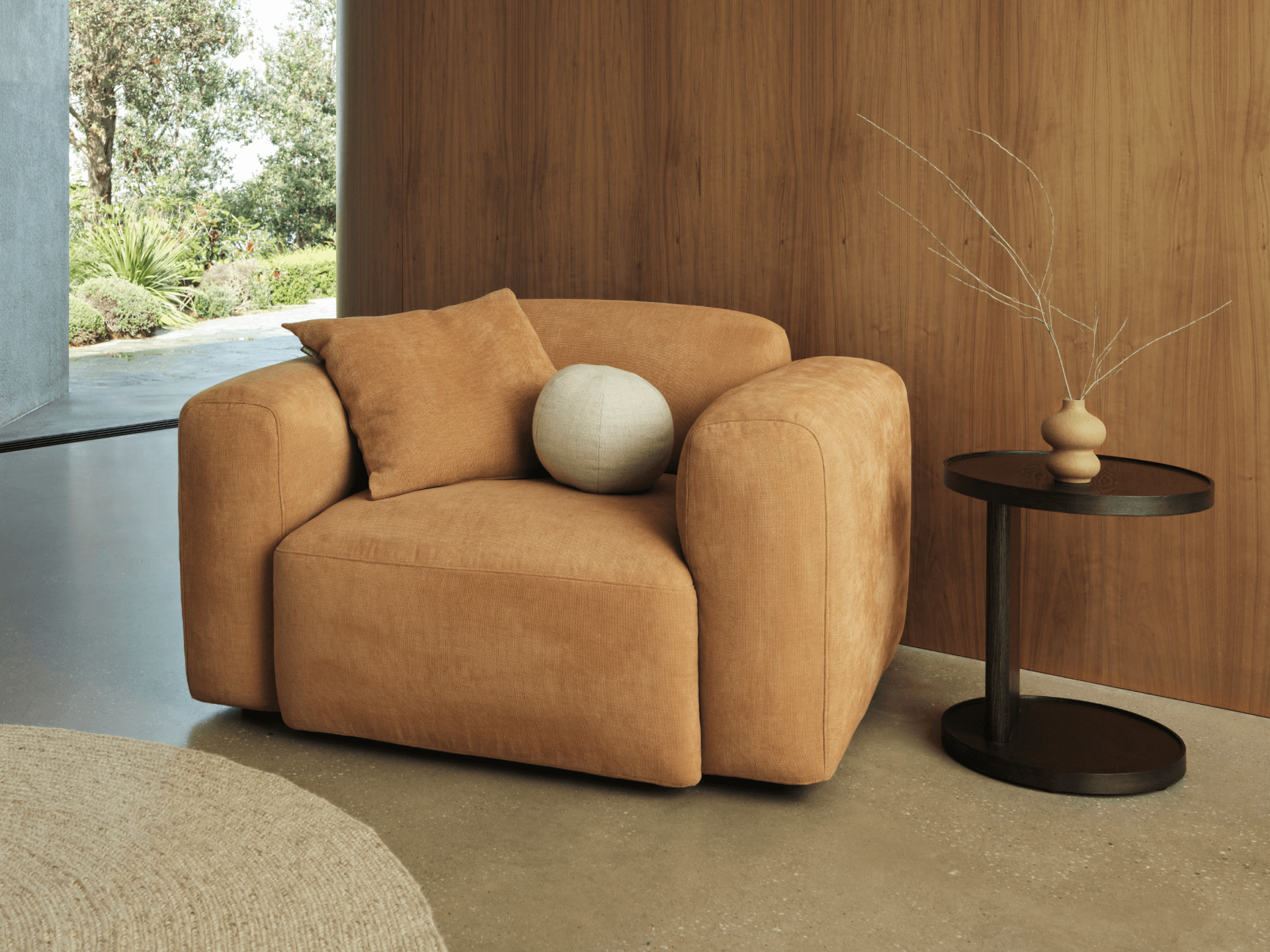 Plush tan armchair in soft suede, featuring clean lines and a cushioned seat. Paired with black round side table, set against warm wood panelling.