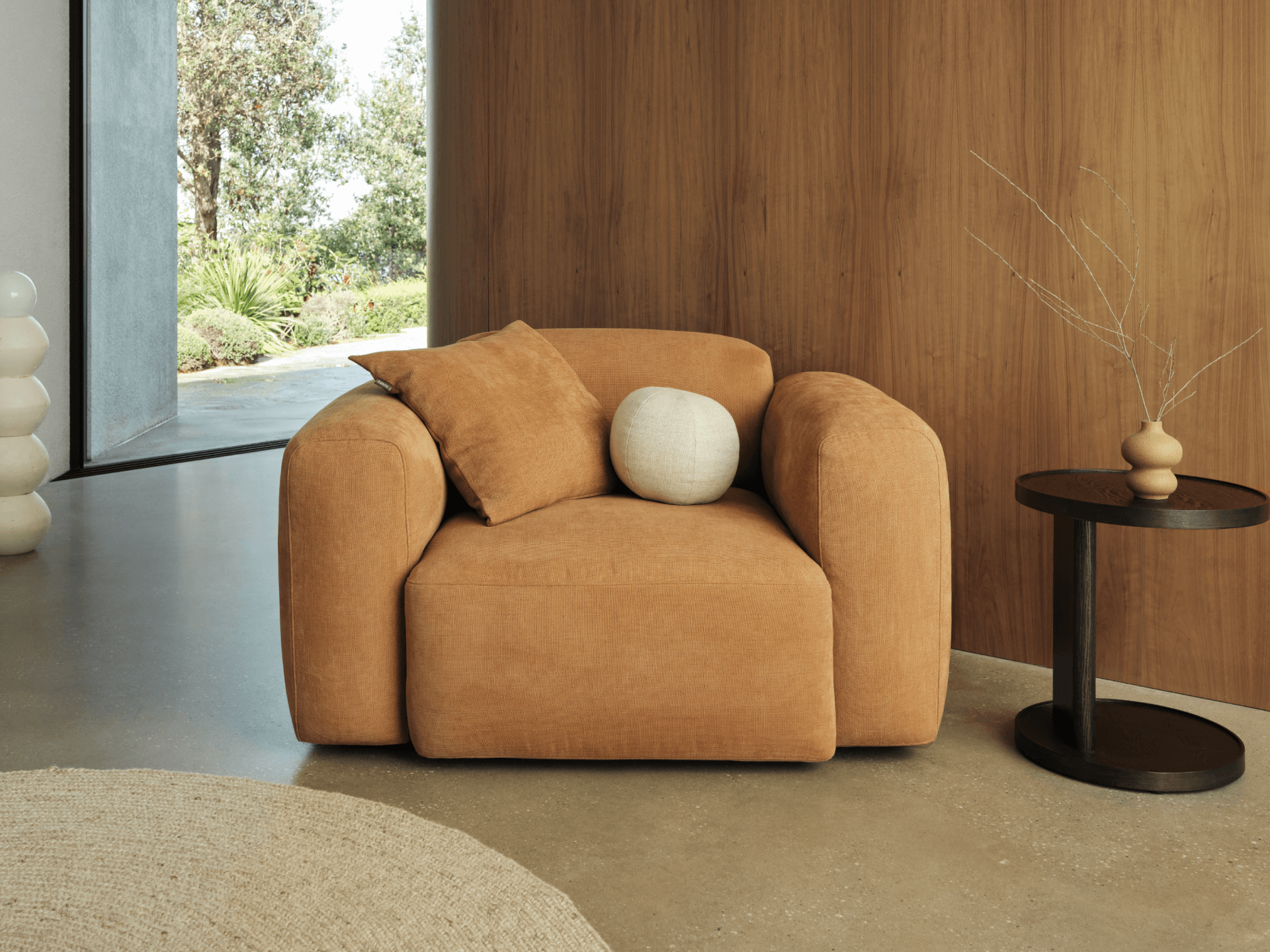 Plush terracotta upholstered armchair with rounded arms, complemented by soft cushions. Set in a modern room with concrete floors and wood panelling.