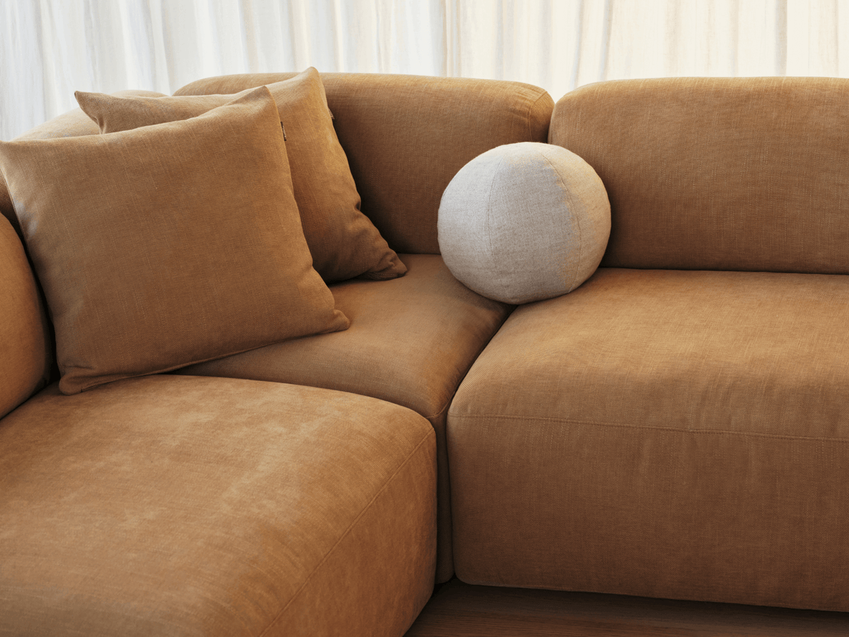 Plush sectional sofa in warm tan velvet, featuring deep cushions and matching pillows. Set in a softly lit modern living space with sheer curtains.