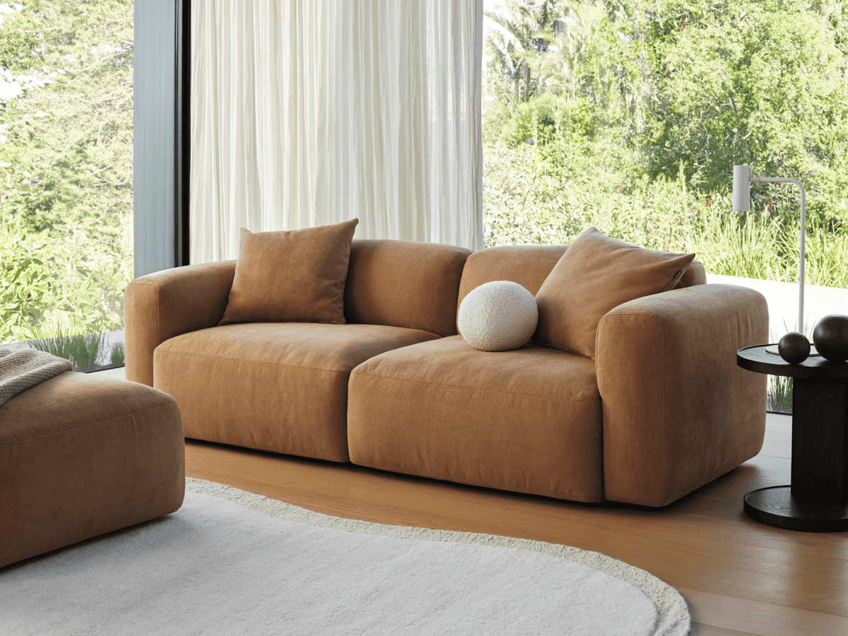 Luxurious brown suede sofa with plush, deep cushions and rounded arms, set on hardwood flooring against a backdrop of sheer white curtains and greenery.