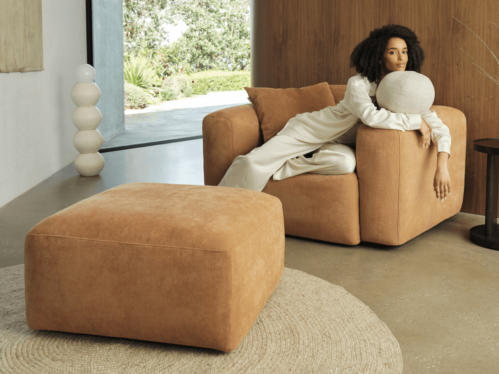 Plush terracotta lounge chair and matching ottoman in soft suede, featuring clean lines in a sunlit, modern living space with concrete floors and wood accents.