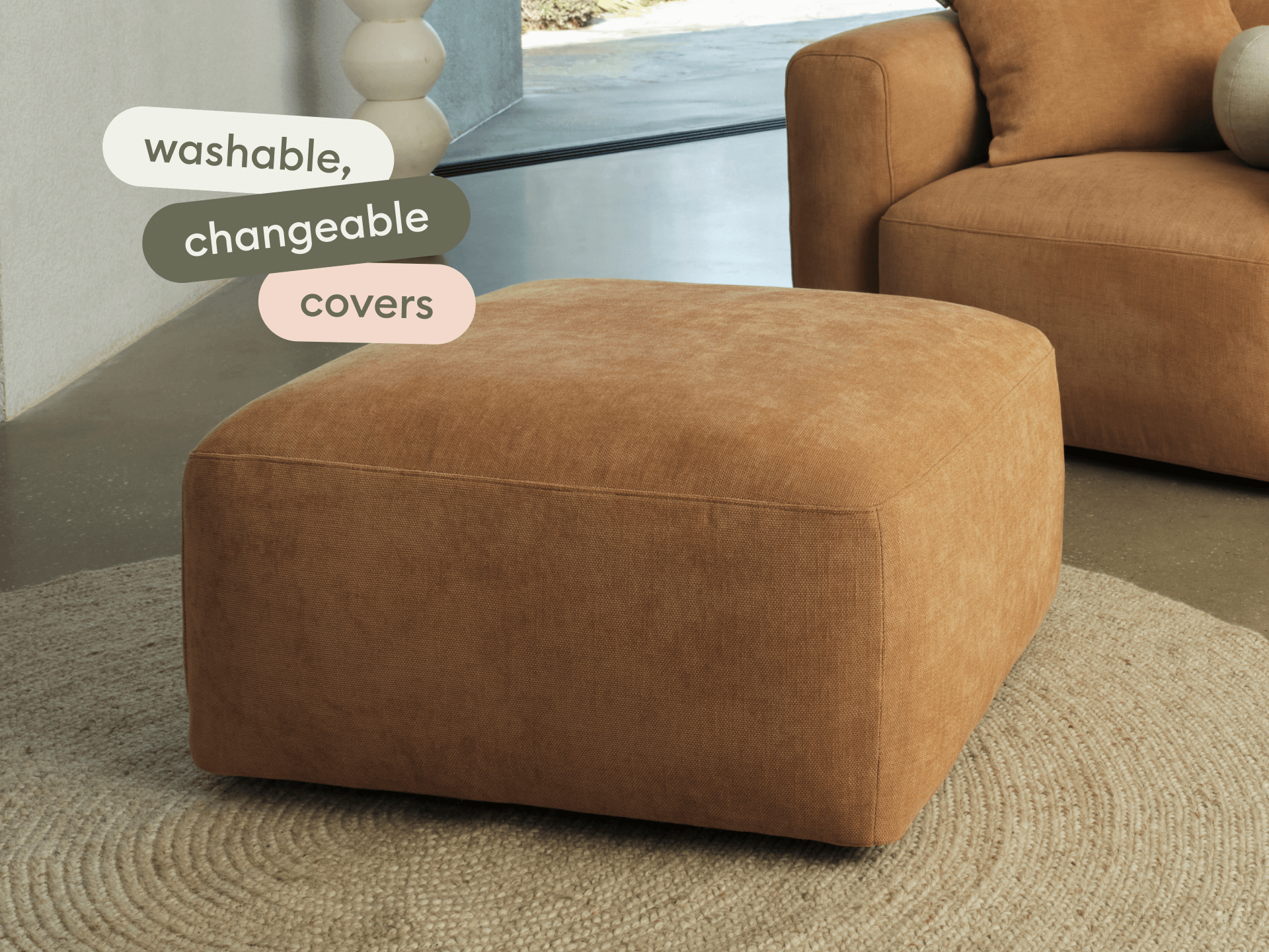 Large square ottoman in rich, caramel fabric with washable covers, set on textured beige rug. Modern living room with concrete floors.