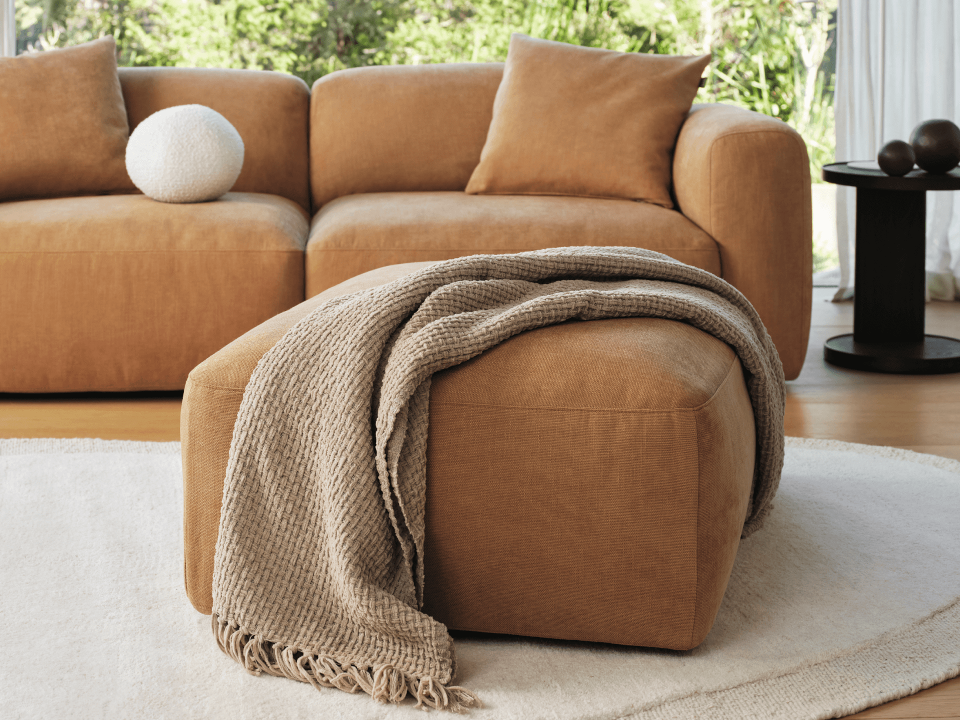 Tan fabric ottoman with soft upholstery, featuring clean lines and plush design. Complements a modern lounge setting, accented by a textured throw.