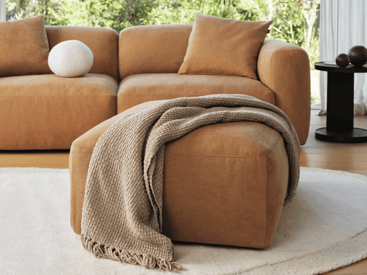 Tan fabric ottoman with soft upholstery, featuring clean lines and plush design. Complements a modern lounge setting, accented by a textured throw.