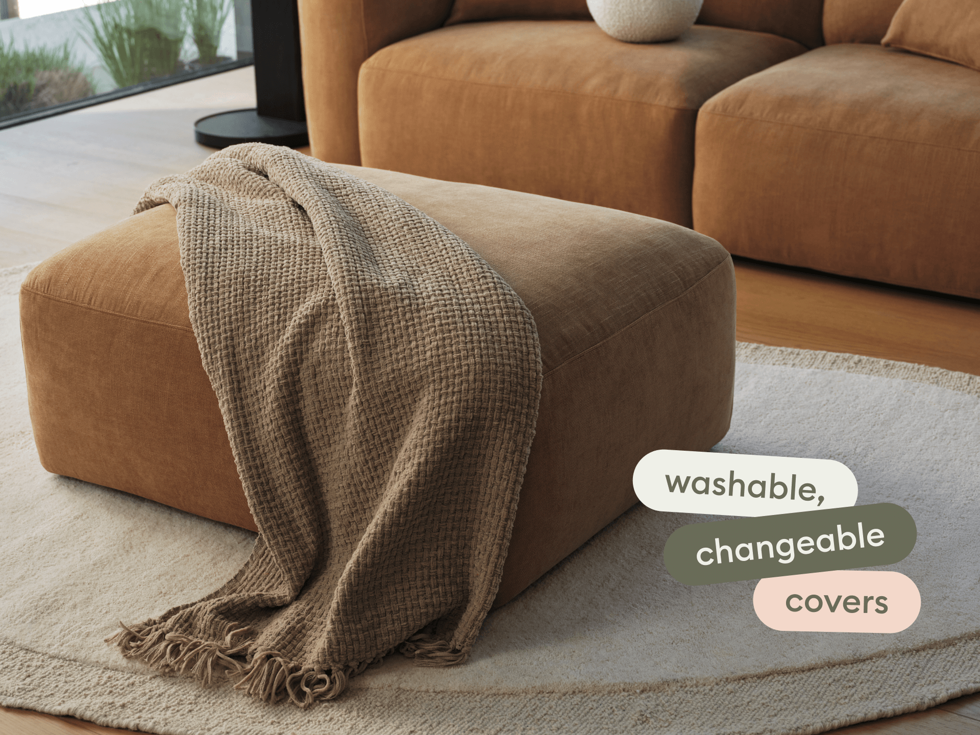 Elegant brown suede ottoman with a plush texture, highlighted by a neutral throw. Set on a light rug in a sleek, modern living room.