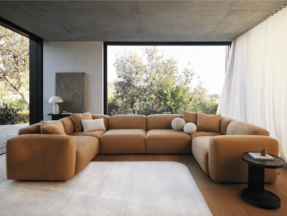Luxurious tan suede modular sofa with plush cushions, set in a modern living room with floor-to-ceiling windows and minimalist decor.