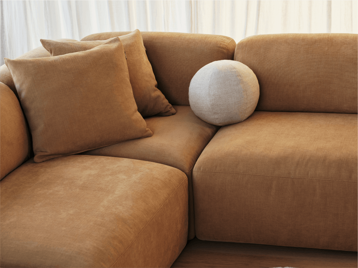 Luxurious brown fabric sectional sofa with plush cushions and a white round pillow. Features modern curved design. Set in a softly lit room.