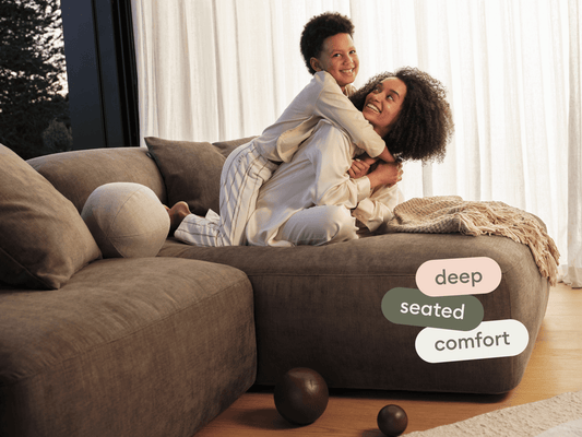 Luxurious grey sectional sofa, plush fabric, deep seats, set in a sunlit living area with natural wood floors and sheer white curtains.