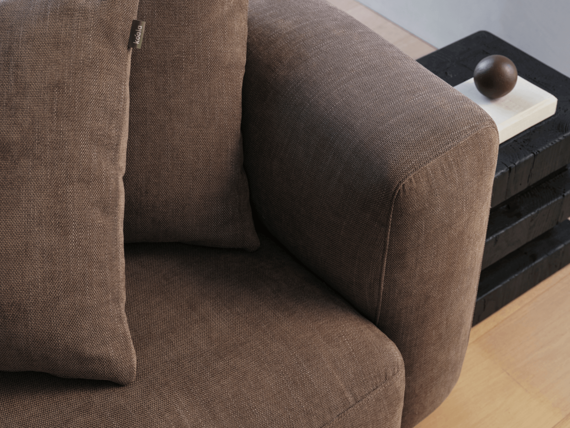 Brown fabric armchair with plush cushions, showcasing soft linen texture. Set against a minimalist light wood floor and stacked black side table with a decorative orb.