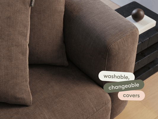 Brown fabric armchair with washable, changeable covers, featuring plush cushions. Set in a modern interior with wooden flooring and a sleek black side table.