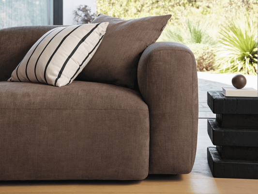 Luxurious brown linen sofa with plush cushioning, complemented by a striped pillow. Situated on a light wood floor beside a modern, stacked black side table.