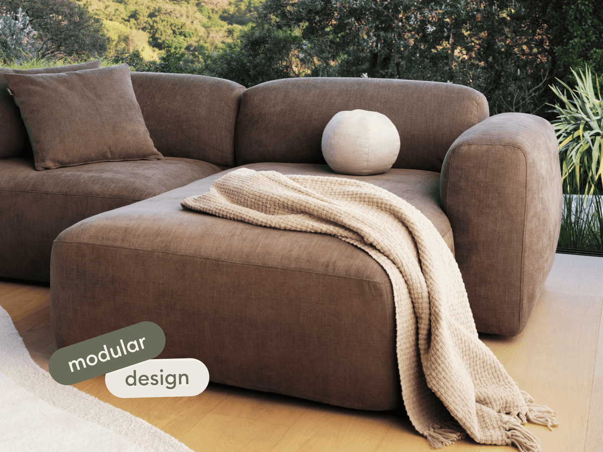 Brown suede modular sofa with plush cushions and a beige knitted throw, set on light wooden flooring amidst lush greenery views in a serene setting.