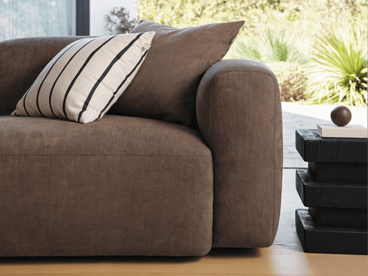 Luxurious brown linen sofa with plush cushions, featuring a contemporary design. Positioned in a sunlit room, paired with a modern black side table.
