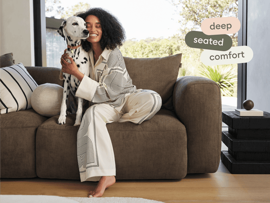 Luxurious deep brown sofa with plush fabric and oversized cushions, set in a sunlit living room with modern decor and large glass windows.