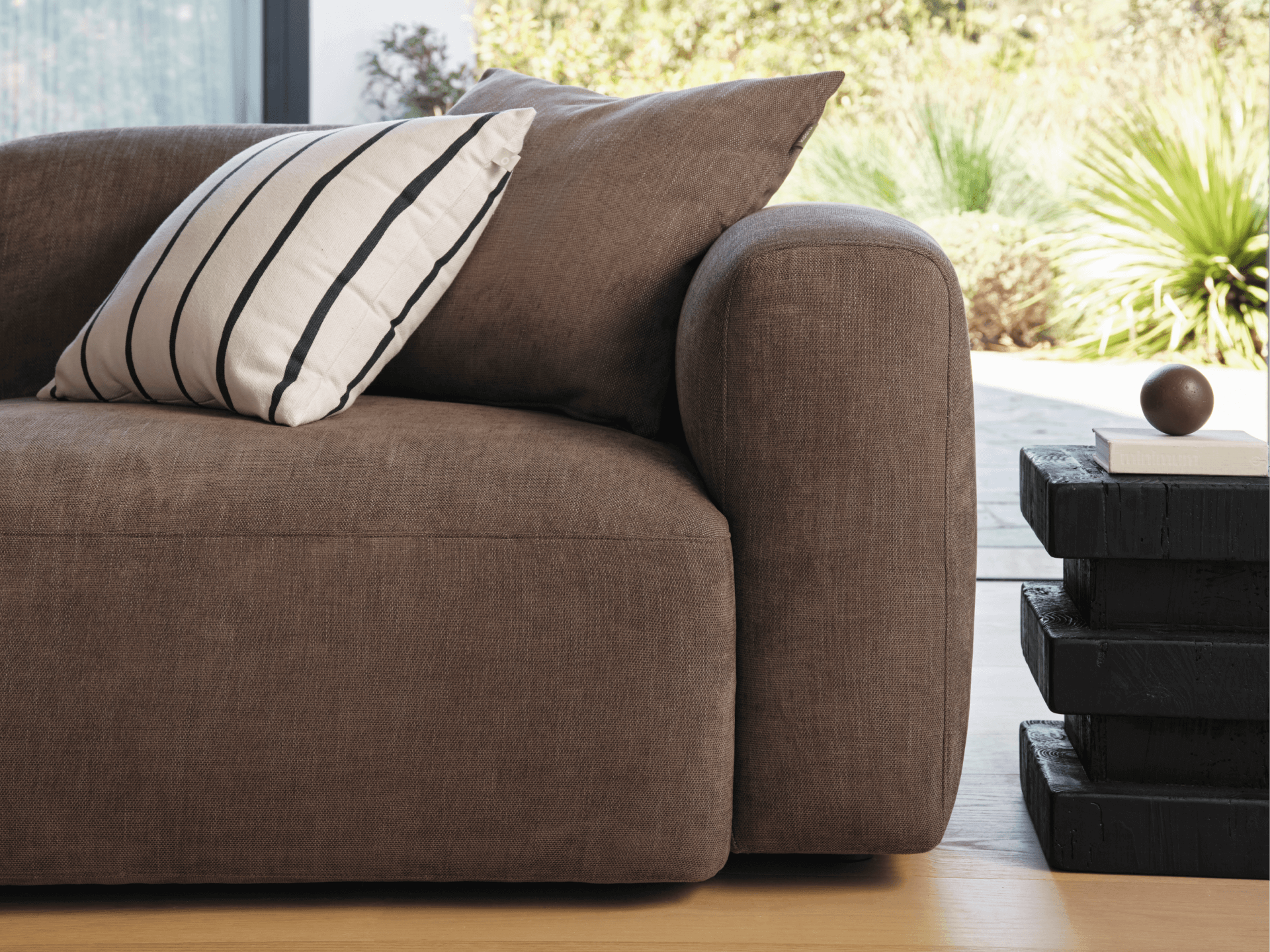 Luxurious brown linen sofa featuring plush cushions, set in a sunlit room with a minimalist black side table and a striped accent pillow.