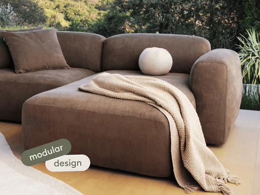 Modular brown fabric sofa with plush cushions, featuring a chaise lounge. Set on a light wood floor, it offers modern elegance ideal for luxury living spaces.