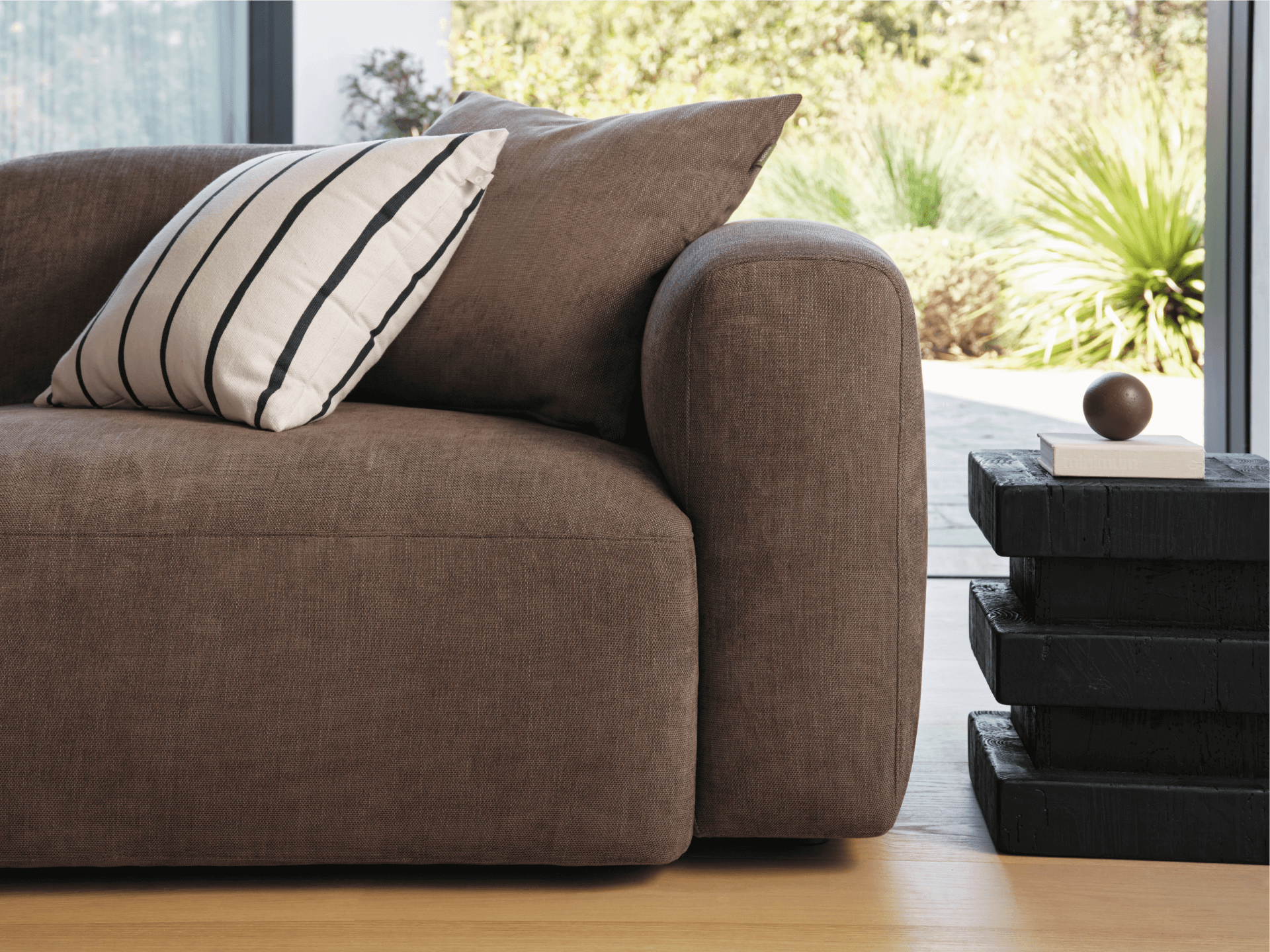 Luxurious brown linen sofa with rounded arms, paired with a striped cushion. Set on light wooden flooring, adjacent to a sleek black side table.