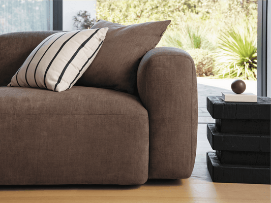 Luxurious brown linen sofa with rounded arms, paired with a striped cushion. Set on light wooden flooring, adjacent to a sleek black side table.