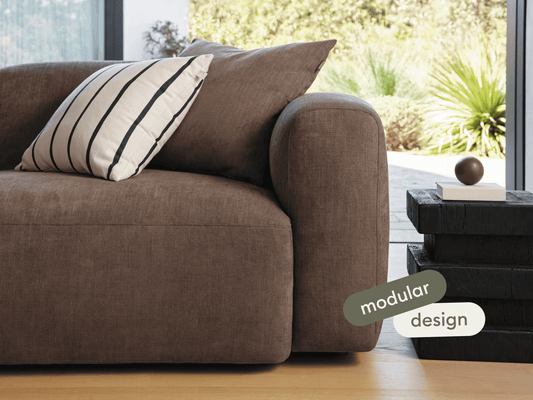Luxurious brown fabric modular sofa with plush cushions. Features rounded arms and sleek lines. Sits in a sunlit room with a striped cushion and dark side table.