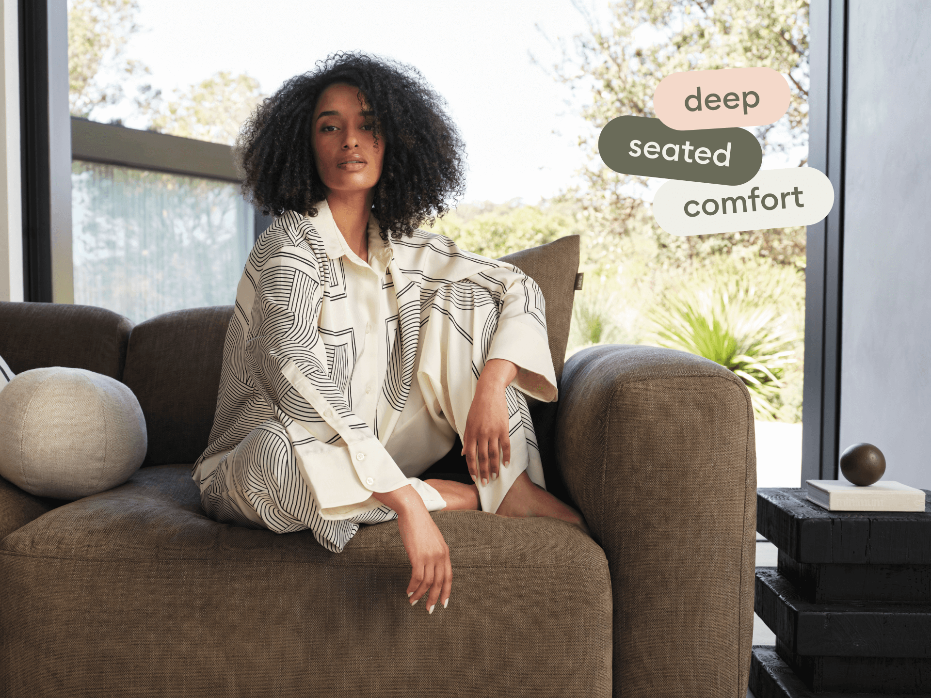 Luxurious deep-seated sofa in rich brown fabric, showcasing plush cushions and wide armrests. Set in a bright, modern living space with garden views.