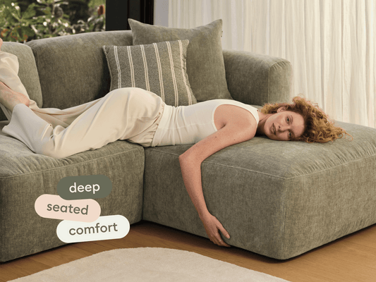 Luxurious deep-seated sofa in muted green fabric, plush cushions, and minimalist design; set on light timber flooring with airy, elegant drapery.