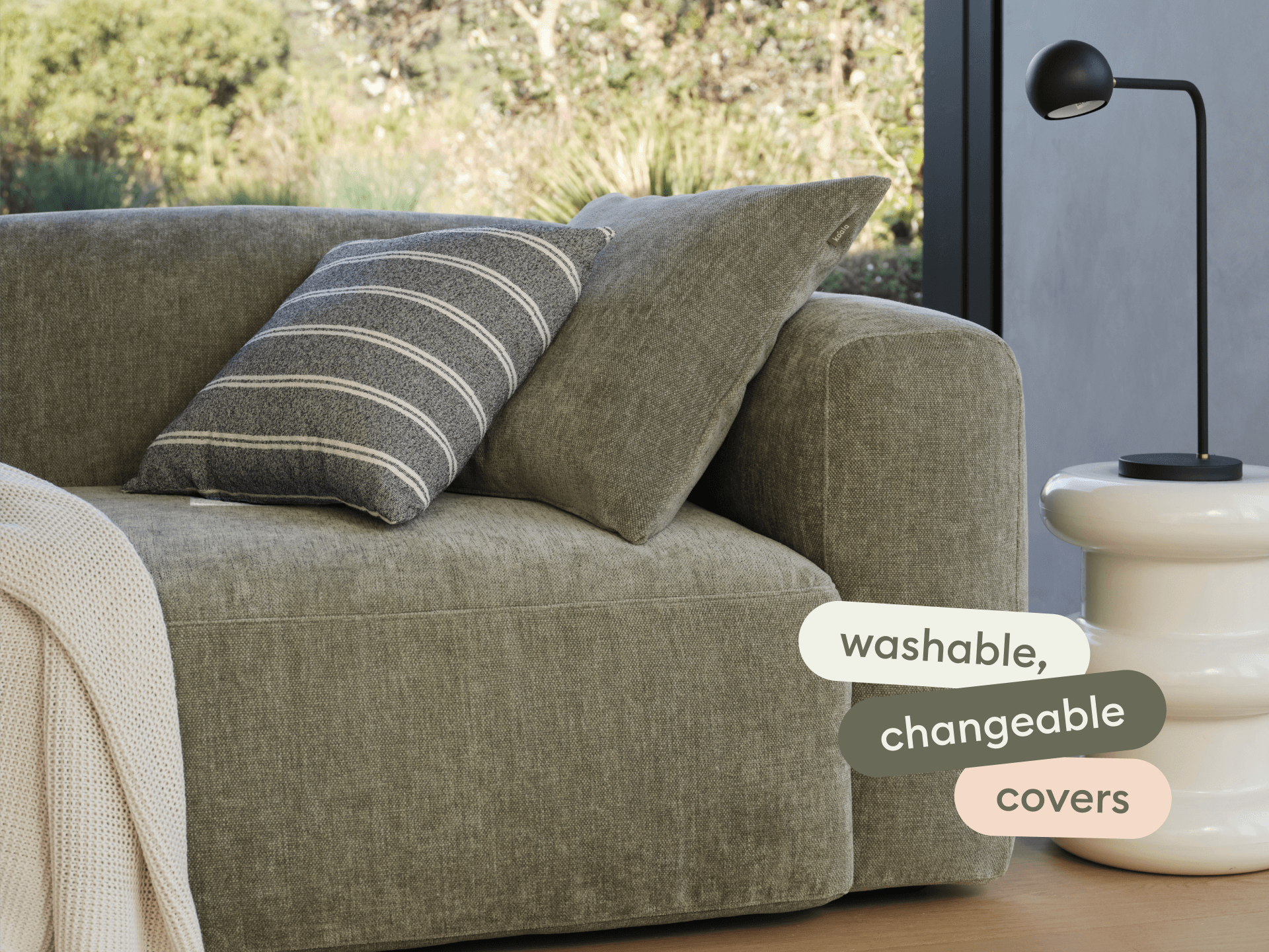 Premium olive green sofa with plush, washable covers and a striped cushion. Set beside a sleek white side table and lamp in a sunlit room.