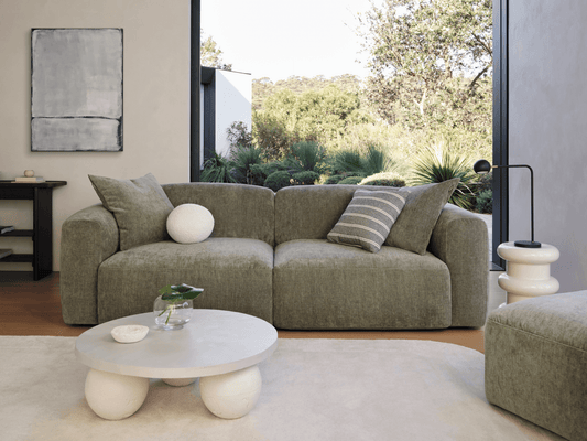 Luxurious olive green linen sofa features deep cushioning and sleek lines, set in a minimalist living room with neutral tones and expansive garden views.