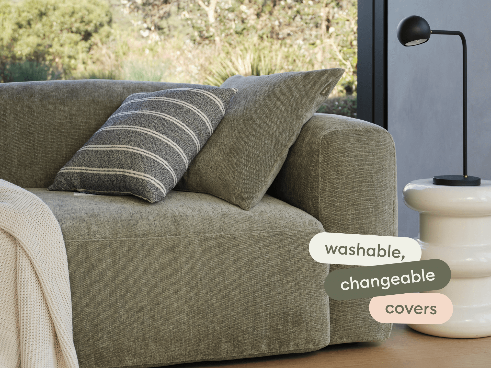 Luxurious olive green sofa with plush, washable linen covers. Features boxy design and a striped cushion. Paired with a chic white side table and sleek lamp.
