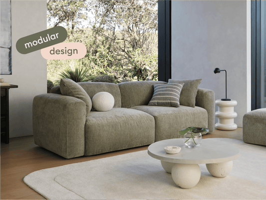 Luxurious modular sofa in olive-green fabric, featuring plush cushions and wide armrests. Set in a sunlit living room with floor-to-ceiling windows.