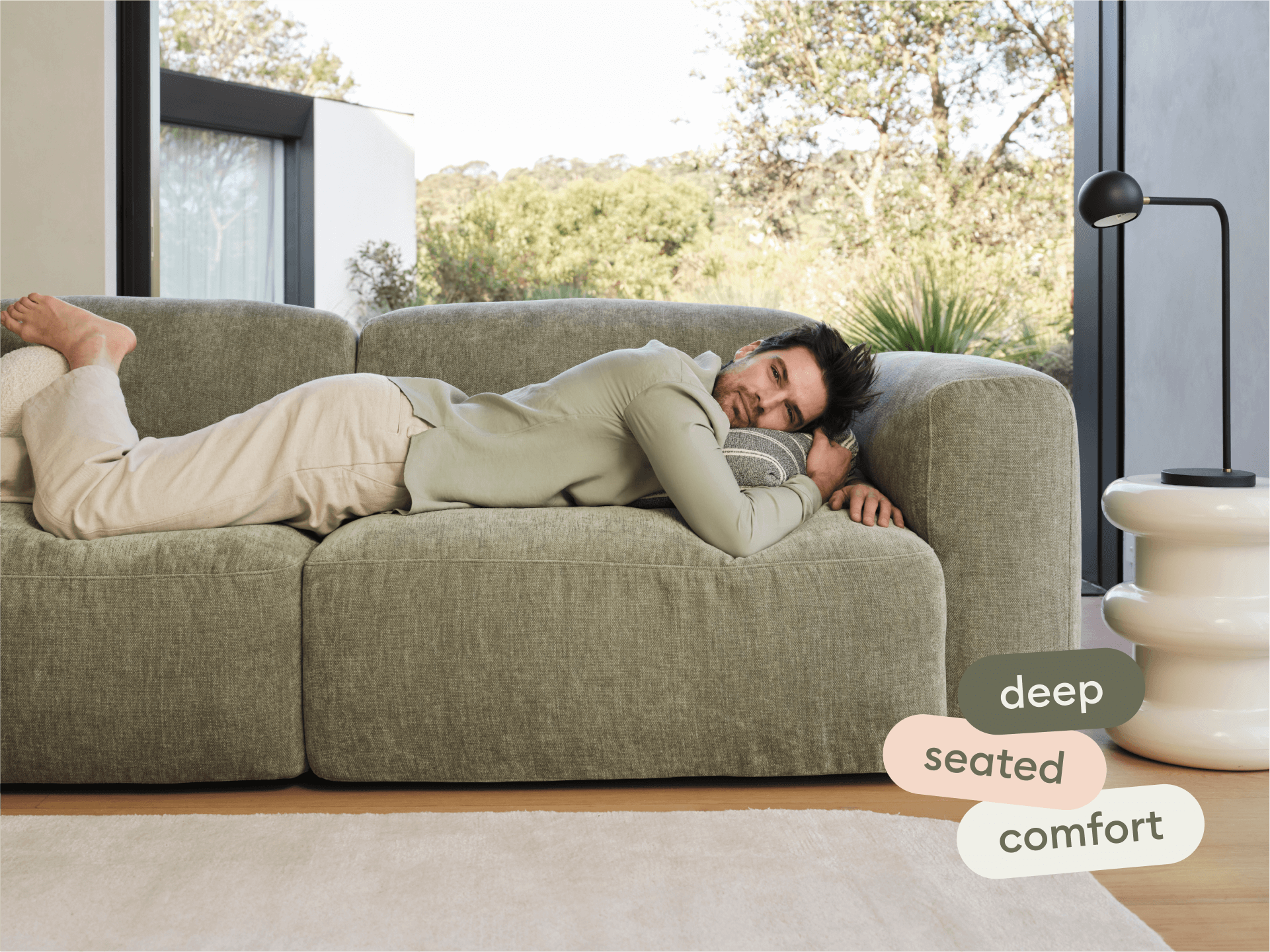 Luxurious olive-green linen sofa with deep seating, rounded edges, nestled in a light-filled room. Features a modern white side table and sleek black lamp.