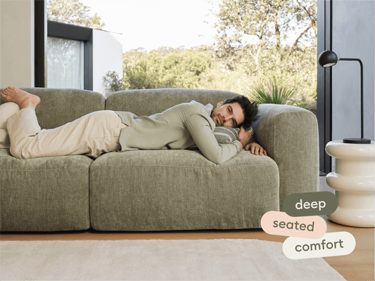 Luxurious olive-green linen sofa with deep seating, rounded edges, nestled in a light-filled room. Features a modern white side table and sleek black lamp.
