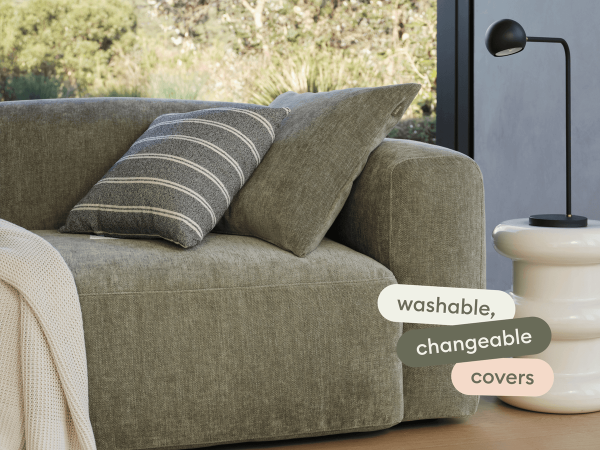 Luxurious olive green fabric sofa with washable, changeable covers. Features a striped cushion. Set in a modern living room with a minimalist side table.