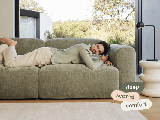 Luxurious deep-seated sofa in soft olive green fabric, featuring plush cushions. Set in a light-filled space with a modern, minimalist side table.