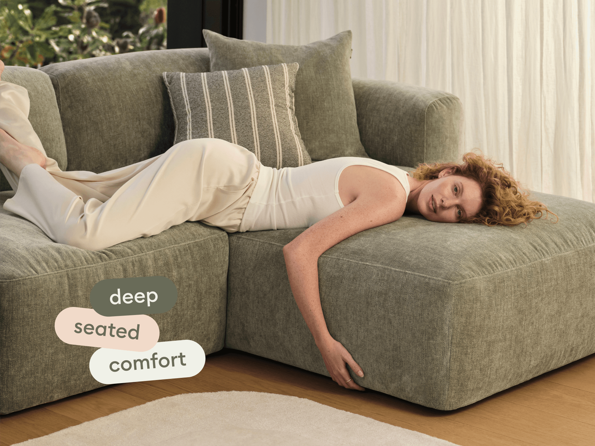 Modern sectional sofa in olive-green fabric, featuring deep seating and plush cushions. Set in a bright room with wooden floors and sheer curtains.