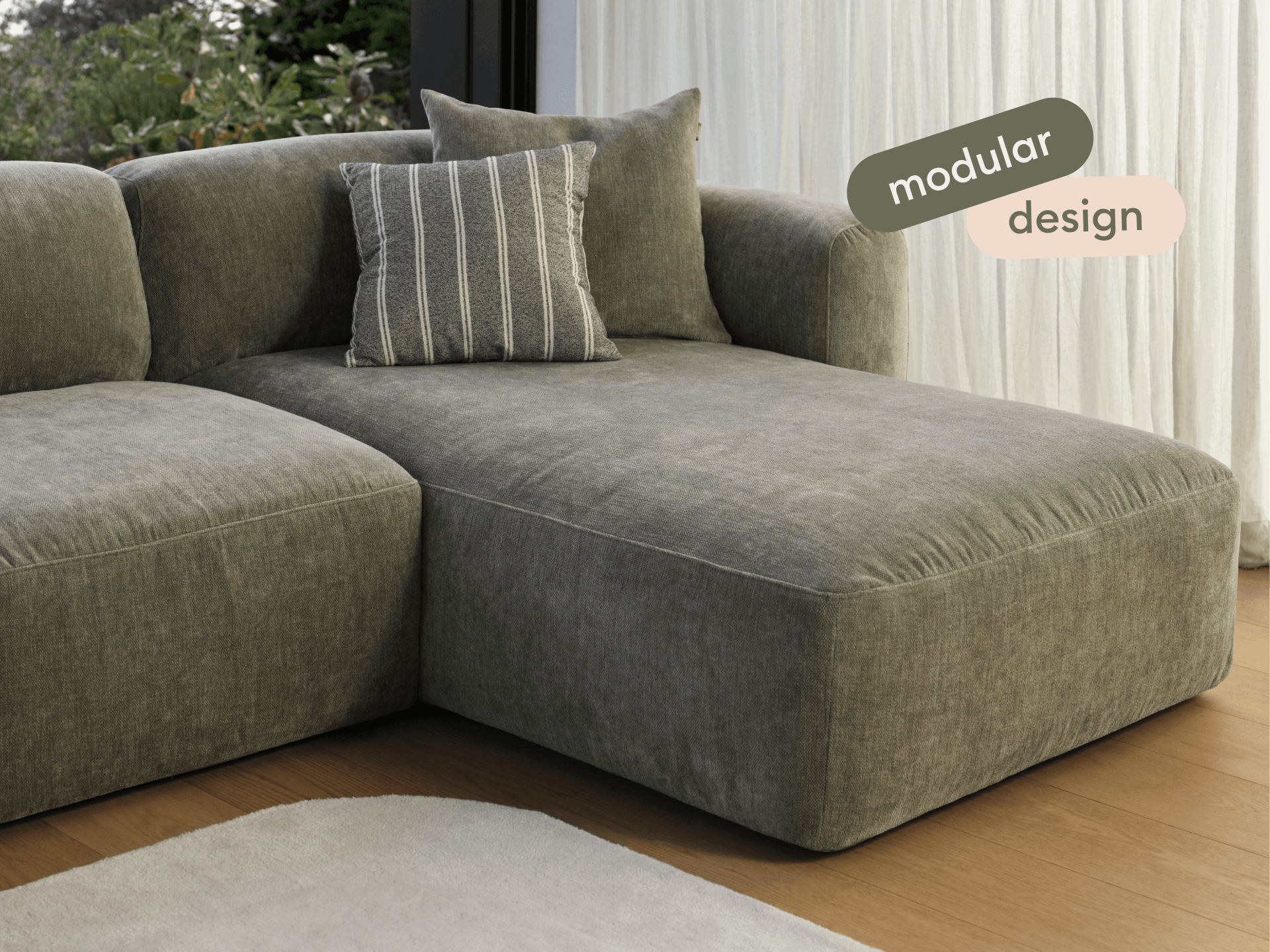 Luxurious modular sofa with plush grey upholstery, featuring a chaise extension. Set on a hardwood floor beside sheer curtains, accent pillow included.