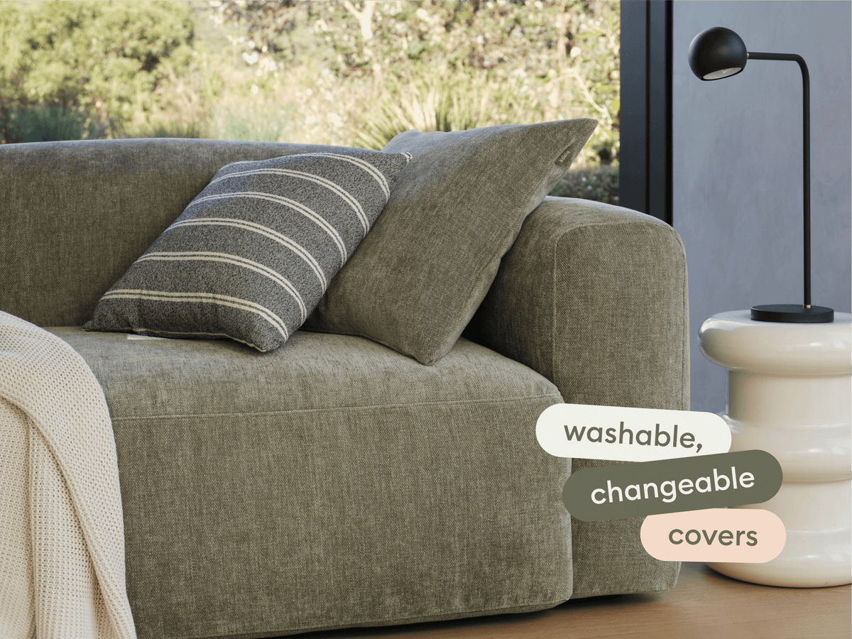 Modern sofa with olive green, washable linen covers and plush cushions. Styled with a striped pillow and woven throw. Set in a bright, minimalist living room.