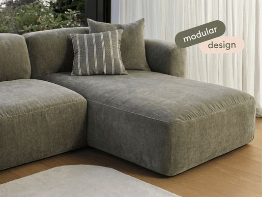 Luxurious grey velvet modular sofa with deep seating, plush cushions, and a sleek design. Set on warm, wooden flooring against sheer white curtains.