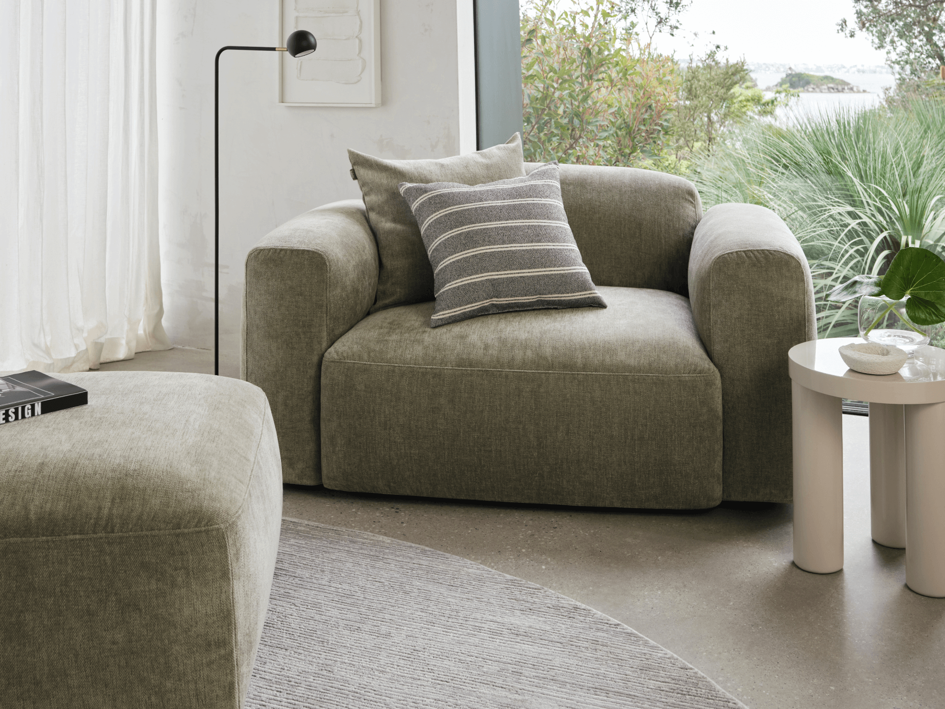 Luxe armchair in plush olive fabric, featuring a wide, low-slung design. Paired with a striped cushion, it sits in a chic, sunlit living space.