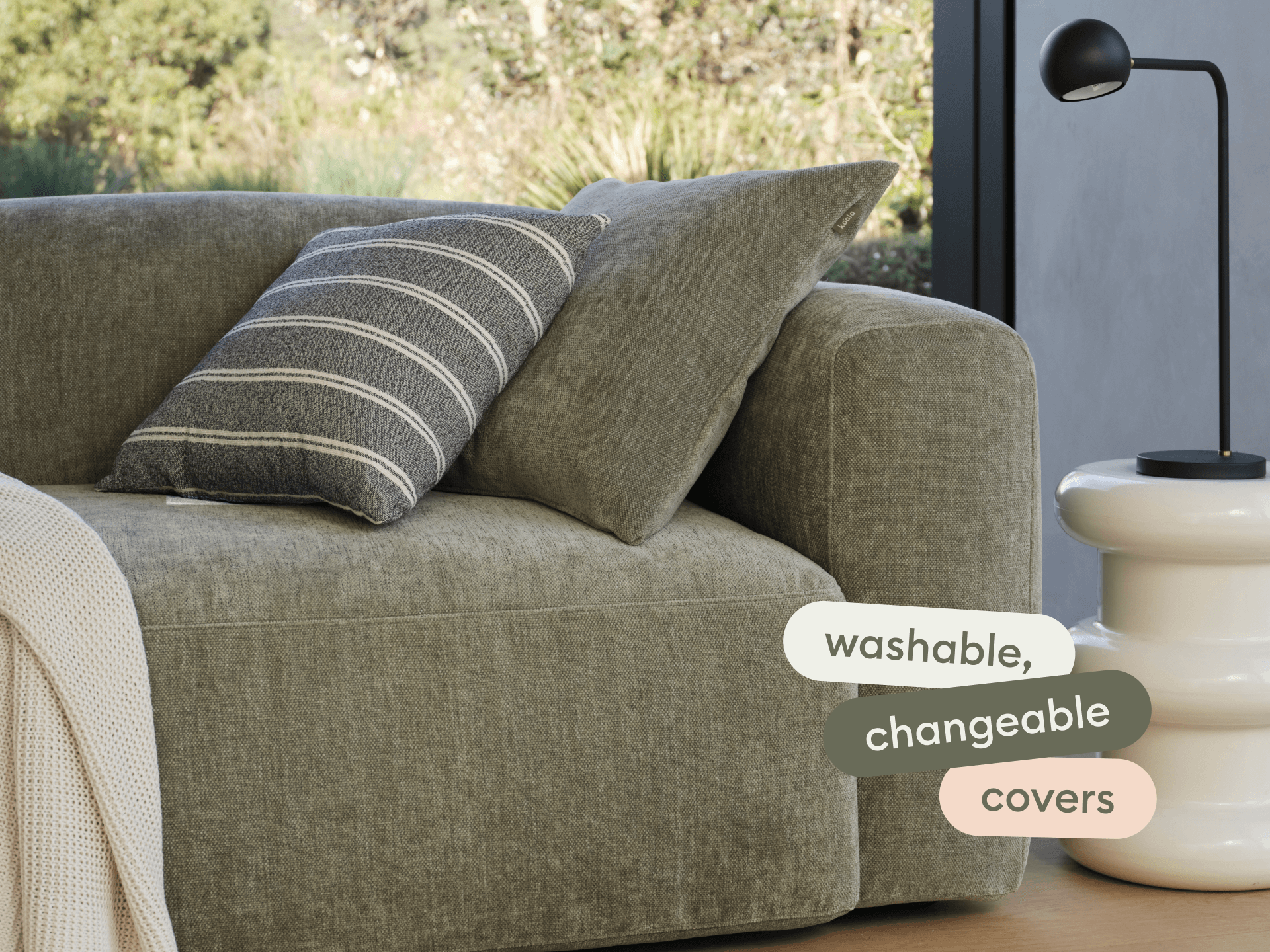 Modern olive green sofa with washable, changeable fabric covers. Features a grey striped cushion. Set beside a minimalist white side table and sleek black lamp.