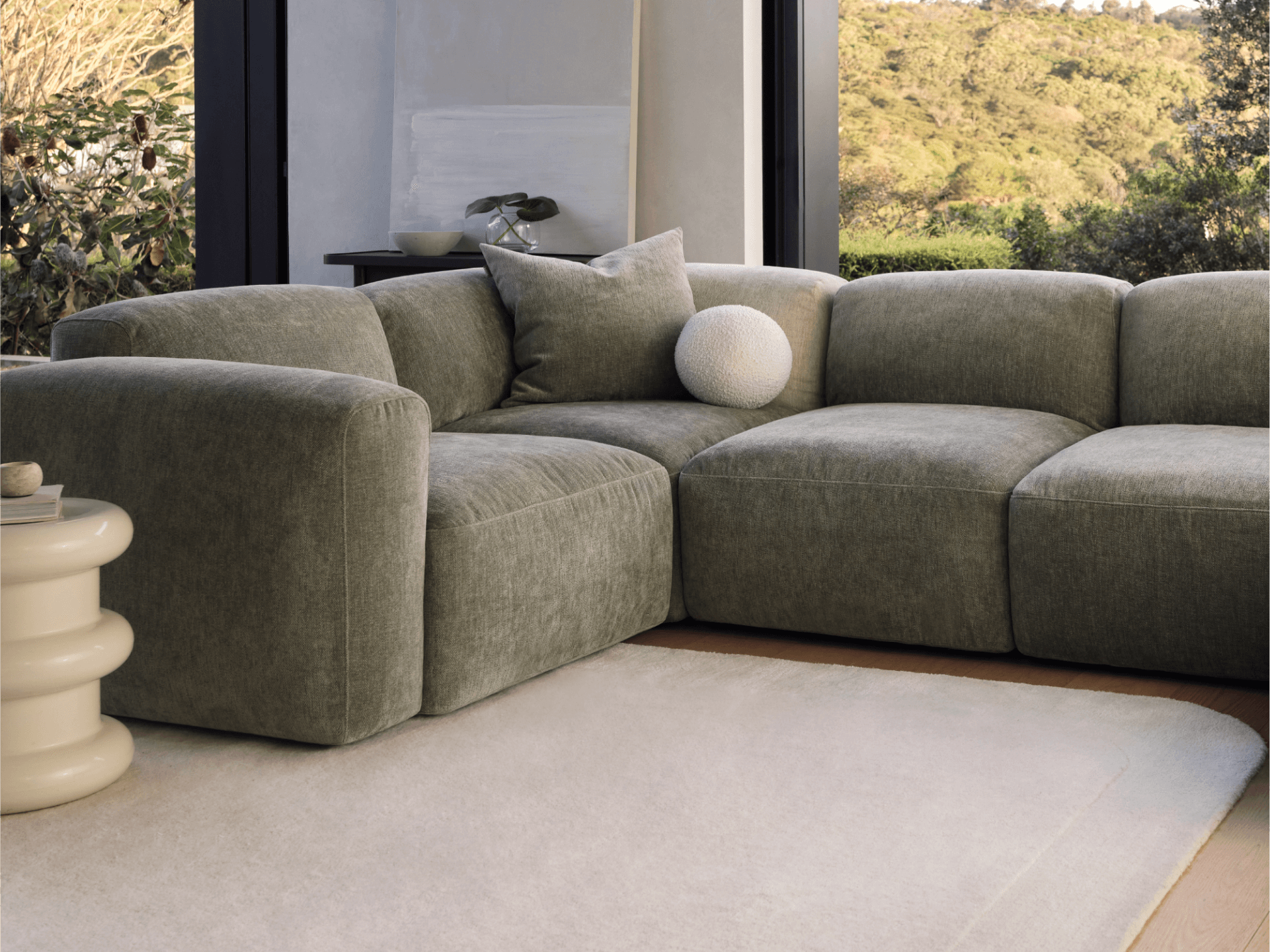 Luxurious grey velvet sectional sofa with plush cushions, modern low-profile design. Set in a light-filled room with scenic views, beige rug, and round side table.